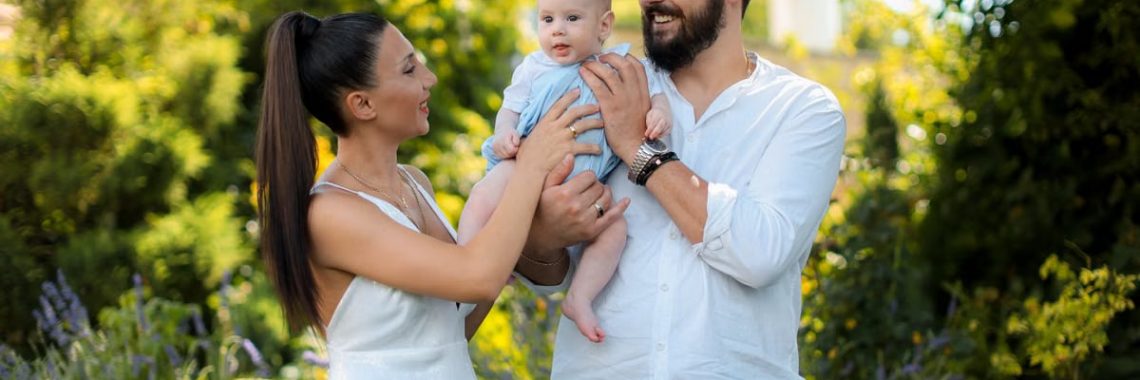 Mother and father joyfully hold their baby in a vibrant outdoor garden setting.