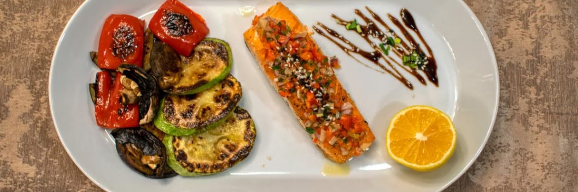 Gourmet grilled salmon served with roasted vegetables and lemon slice on a white plate.