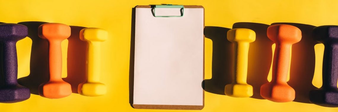 Brightly colored dumbbells arranged with a clipboard on a vibrant yellow background.